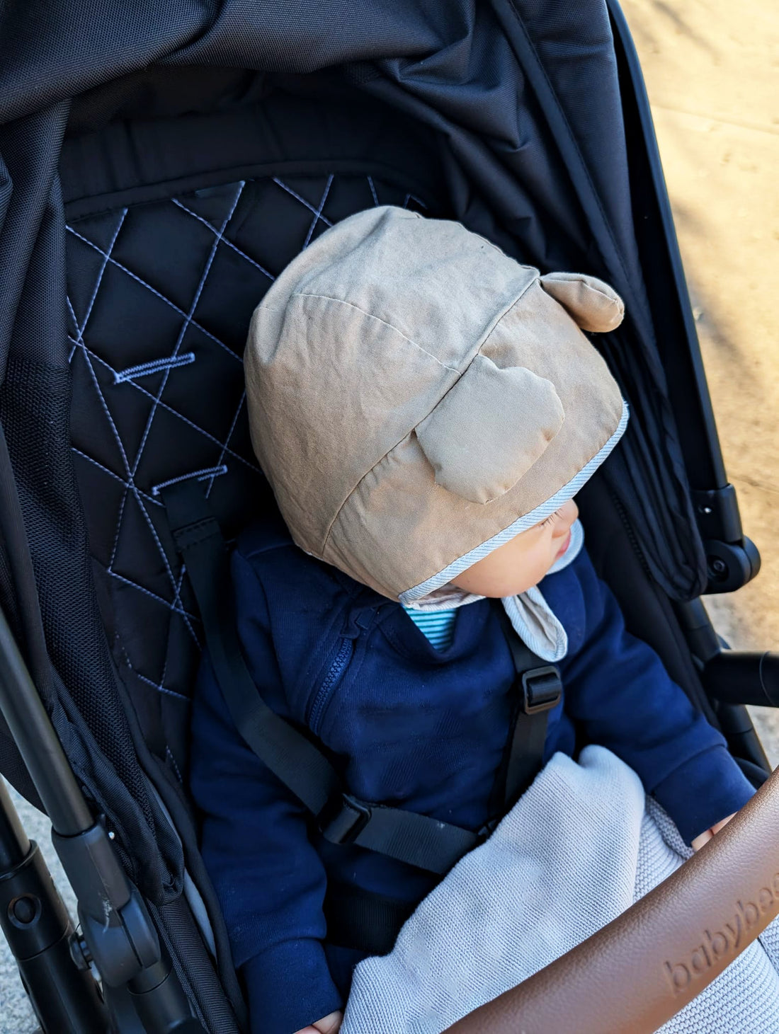 Baby wearing a handmade tan cotton bear bonnet sitting in a pram during a park walk
